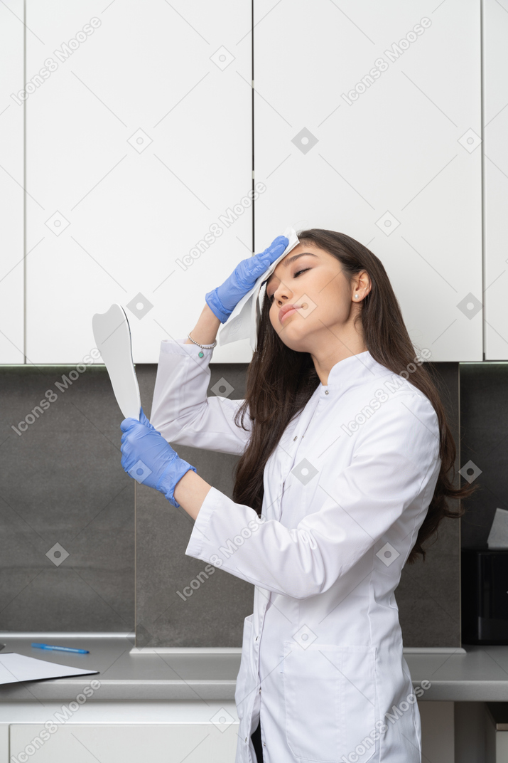 Side view of a female doctor looking in the mirror and wiping her forehead