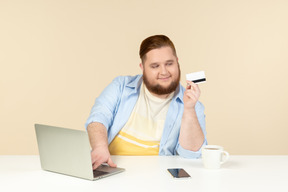 Contento joven con sobrepeso sentado a la mesa y mirando la tarjeta bancaria