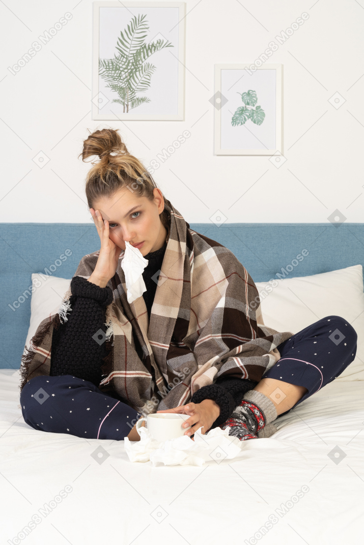 Vista frontal de uma jovem doente de pijama enrolada em um cobertor xadrez na cama com um guardanapo no nariz