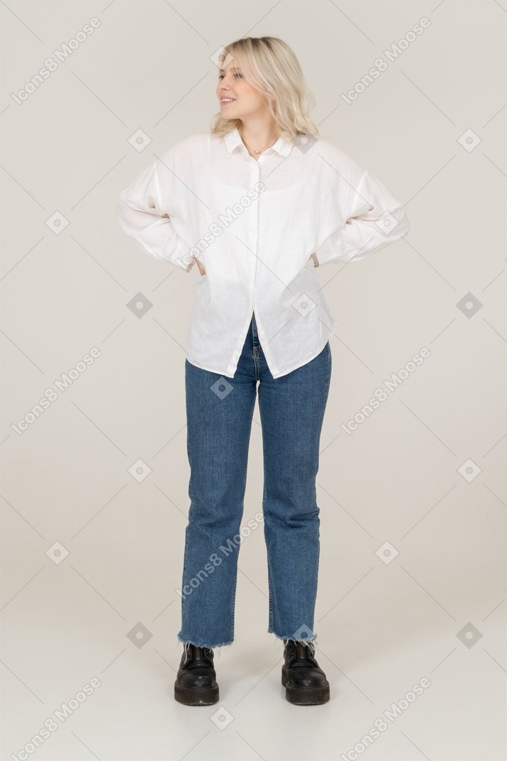 Front view of a blonde female in casual clothes putting hands on hips and looking aside while smiling