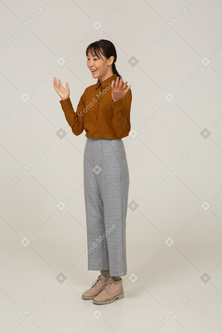 Vista di tre quarti di una giovane donna asiatica felice in calzoni e camicetta che alza le mani