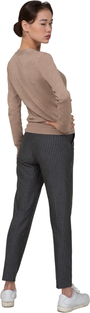 Vista posterior de tres cuartos de una señorita guiñando un ojo en suéter y pantalones poniendo la mano en la cadera