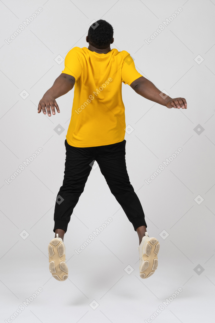 Vista traseira de um jovem de pele escura pulando em uma camiseta amarela estendendo as mãos