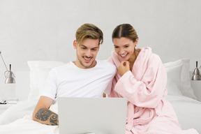 A man and woman sitting on a bed smiling and looking at a laptop