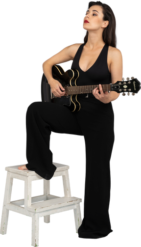 一位年轻的女士，穿着黑色西装，抱着吉他，把腿放在凳子上的四分之三的视图