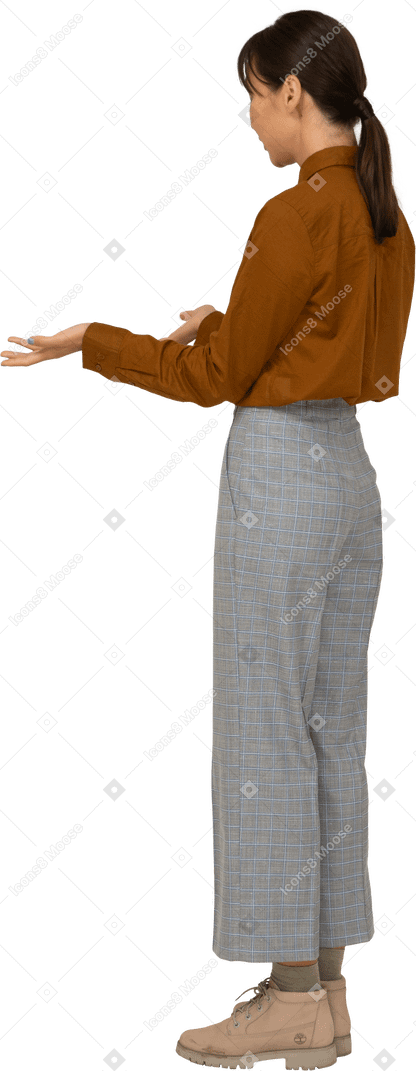 Vista posteriore di tre quarti di una giovane donna asiatica interrogativa in calzoni e camicetta che alza le mani