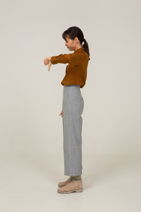 Vista lateral de uma jovem mulher asiática de calça e blusa mostrando o polegar para baixo
