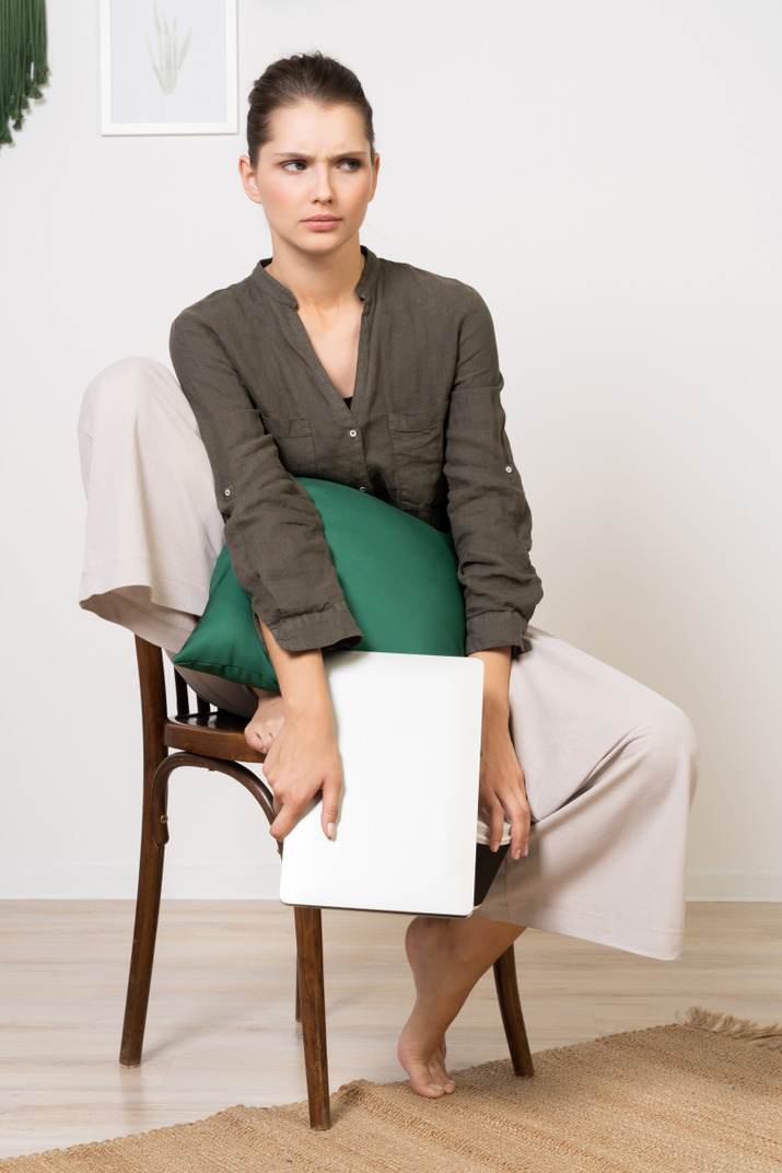 Front view of a confused young woman sitting on a chair and holding her laptop