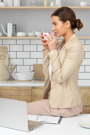 A woman sitting on a kitchen counter drinking a cup of coffee