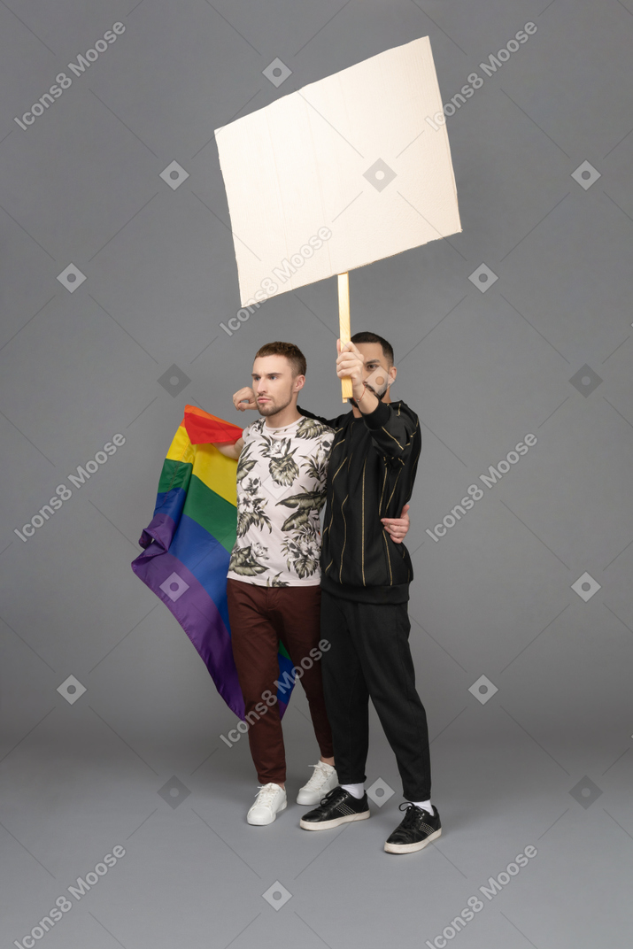 Vista de tres cuartos de dos hombres jóvenes sosteniendo una cartelera y una bandera lgbt