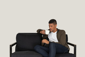 Front view of a young displeased man sitting on a sofa with a cup of coffee