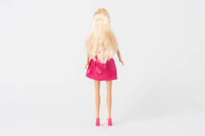 A back shot of a barbie doll in a shiny pink dress and pink high heels standing isolated against a plain white background