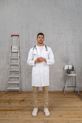 Front view of a young doctor standing in a room with ladder and chair holding hands together