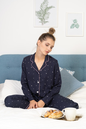 Vue de face d'une jeune femme en pyjama regardant la tasse de café et quelques pâtisseries sur un plateau