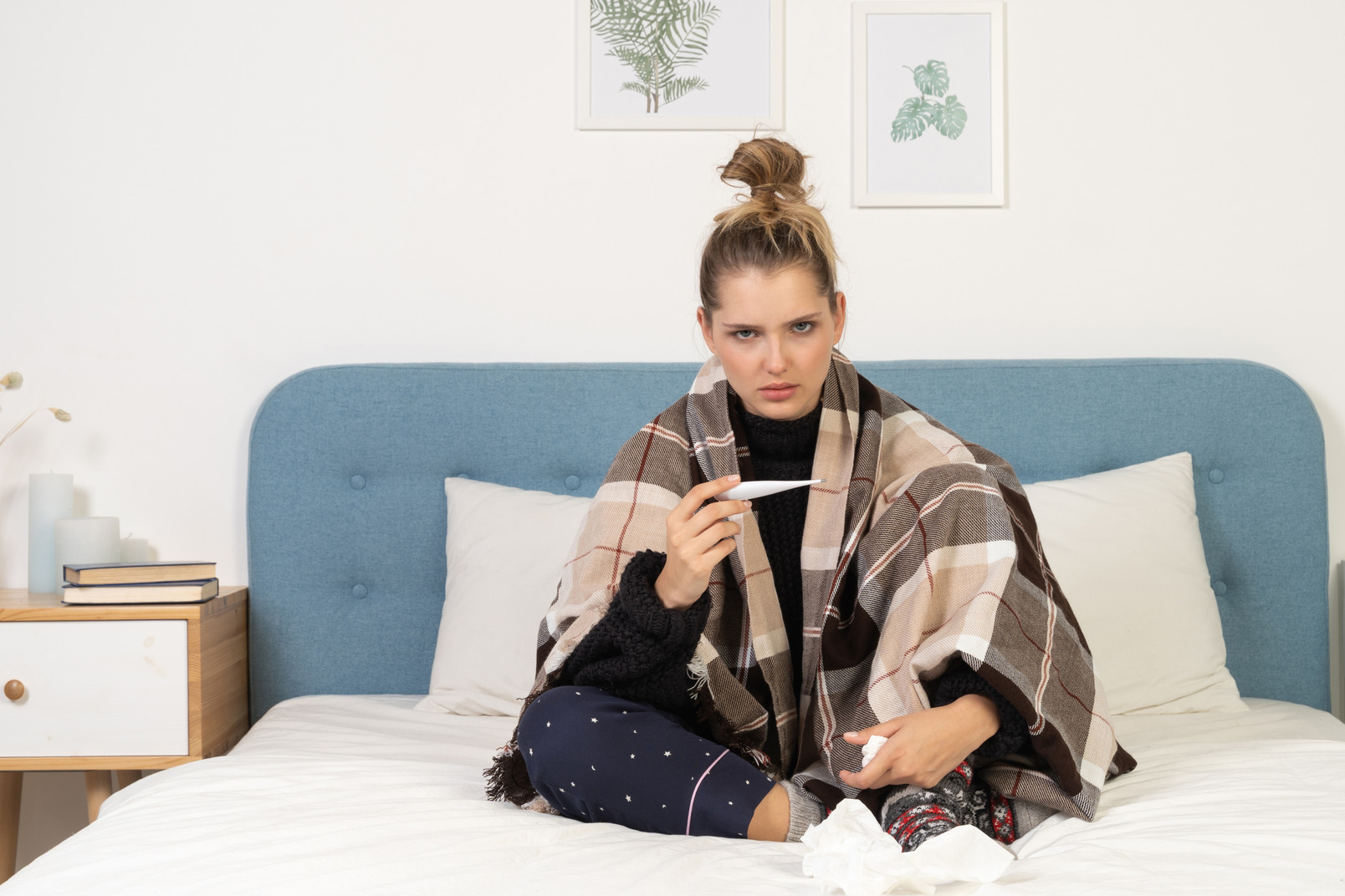 Front view of an angry ill young lady in pajamas wrapped in checked blanket checking her temperature