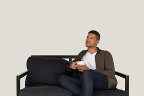 一个年轻的梦想男人坐在沙发上，喝着一杯咖啡的前视图