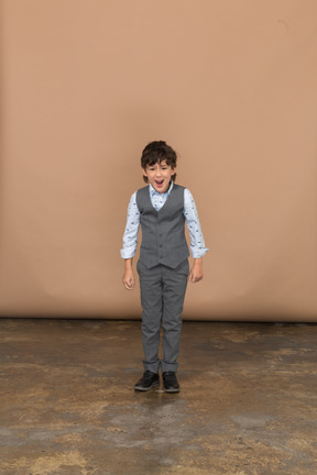 Vista frontal de un niño enojado en traje de pie con los puños cerrados y mirando a la cámara