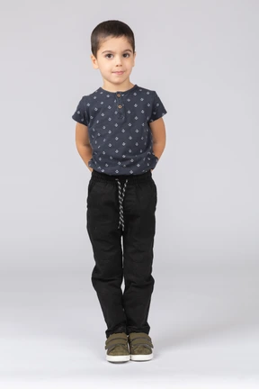 Front view of a cute boy standing with hands behind back and looking at camera