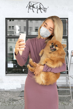 A woman in face mask holding a dog and taking a selfie