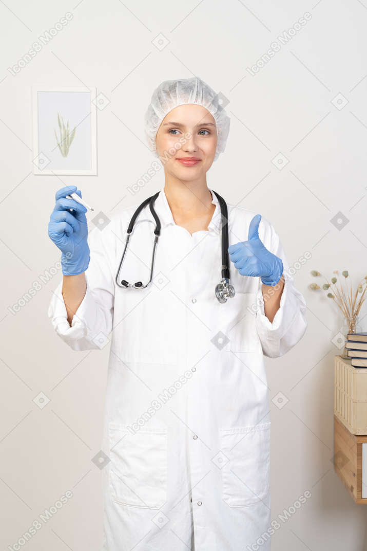 Front view of a young female doctor with stethoscope holding thermometer and showing thumb up