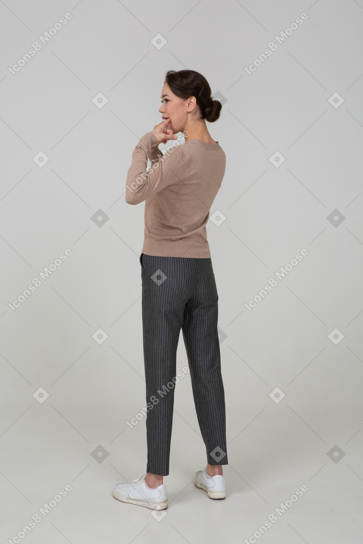 Three-quarter back view of a young lady in pullover and pants touching her mouth