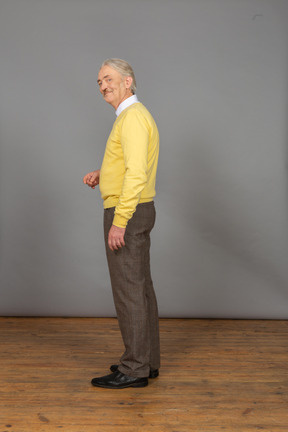 Vista lateral de um velho sorridente de blusa amarela, levantando a mão e olhando para a câmera