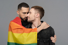 Front view of two young men half-hugging sensually and wearing lgbt flag