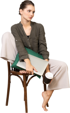 Vue de face d'une jeune femme assise sur une chaise et tenant son ordinateur portable et touchant une tasse de café