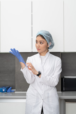 Front view of a young female doctor putting on protective gloves in her medical cabinet