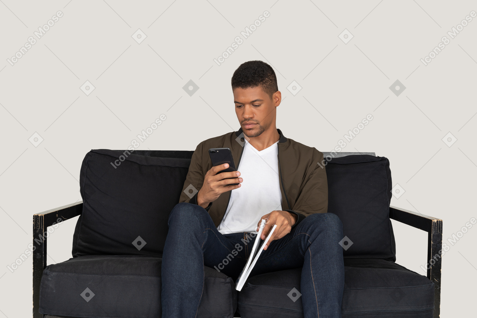 Front view of young man sitting on a sofa holding a magazine while using phone