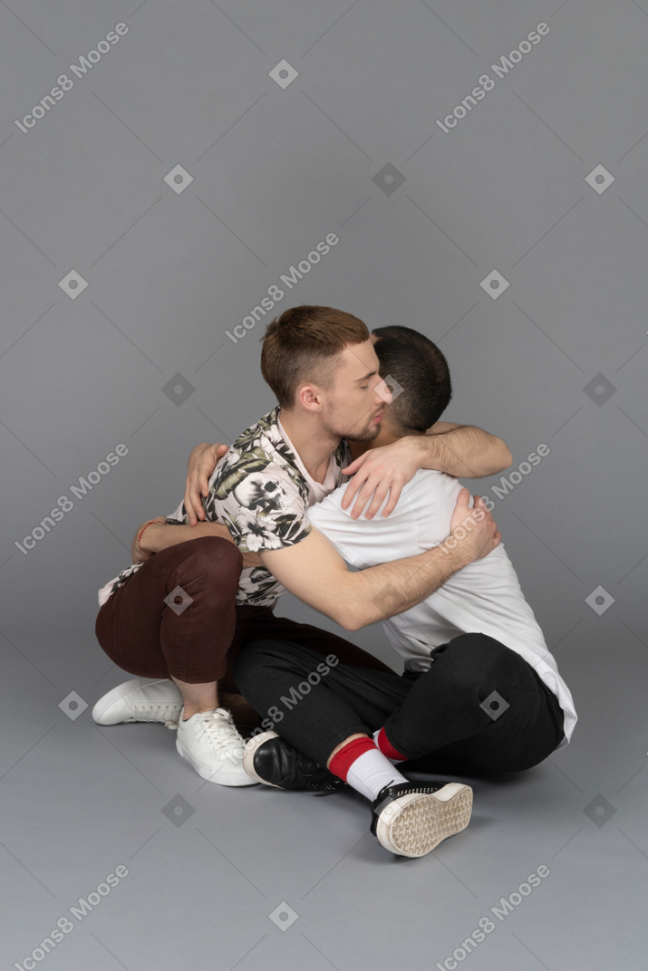 Side view of two young men sitting on the floor and hugging