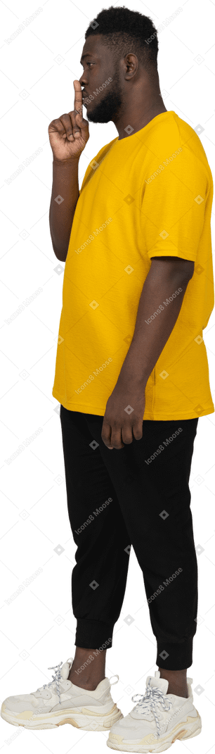 Vista di tre quarti di un giovane uomo dalla pelle scura in maglietta gialla che mostra gesto di silenzio