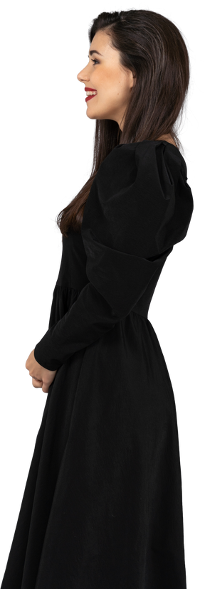Вид сбоку улыбающейся молодой леди в черном платье, стоящей на месте