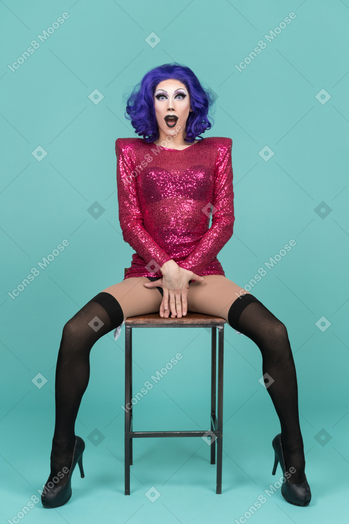 Drag queen cobrindo partes íntimas com as mãos enquanto está sentado em um banquinho