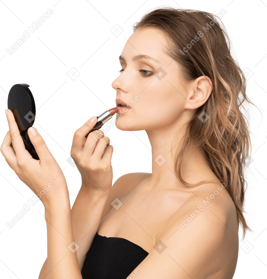 Vista lateral de una mujer joven aplicando lápiz labial mientras sostiene un espejo
