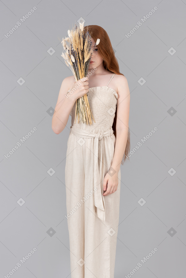 Teenage girl in beige overalls closing half of her face with bouquet of dried flowers