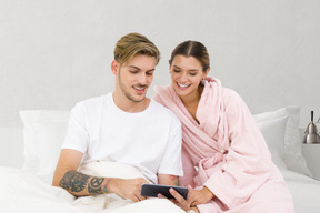 A man and woman sitting on a bed looking at a cell phone