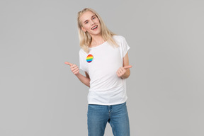 Beau jeune homme aux longs cheveux blonds, debout sur un fond gris, vêtu d'un jean bleu et d'un t-shirt blanc avec écusson lgbt