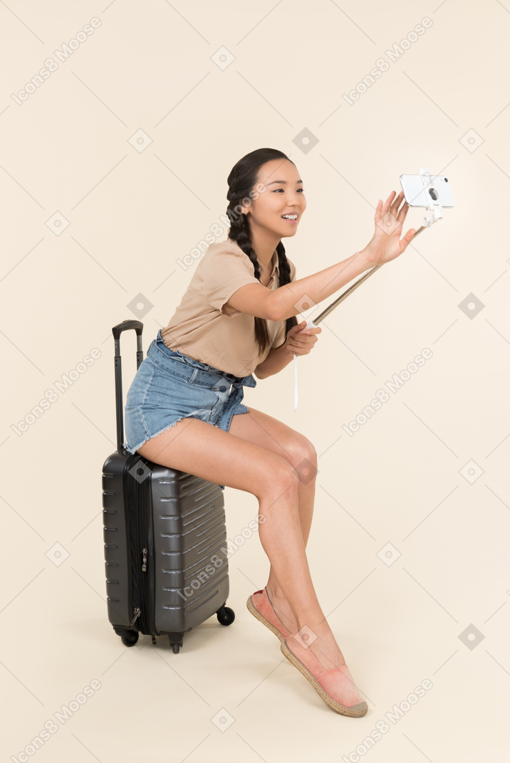 Young female traveller sitting on suitcase and holding selfie stick