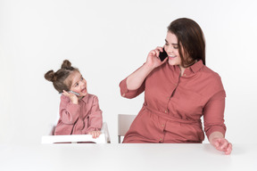 母亲和她的小女儿，穿着红色和粉红色的衣服，坐在餐桌旁，手里拿着智能手机