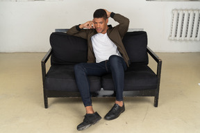 Front view of a perplexed young man sitting on a sofa and talking on his phone