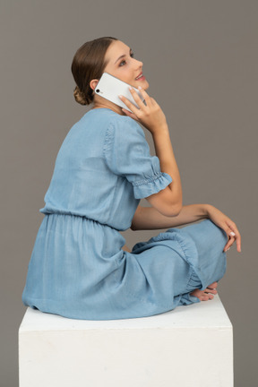 Side-back view of young woman sitting on cube and talking on smartphone