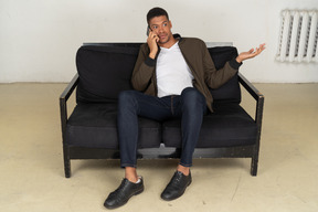 Front view of a perplexed young man sitting on a sofa and talking on his phone