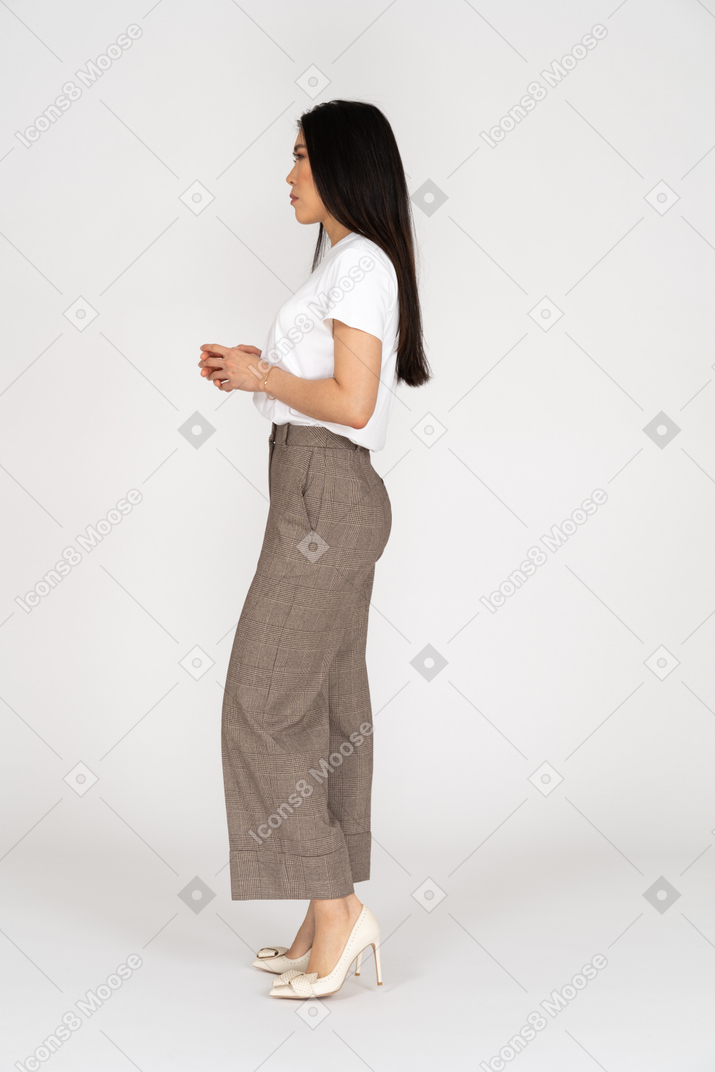 Vista laterale di una giovane donna seria in calzoni e maglietta che tengono le mani insieme