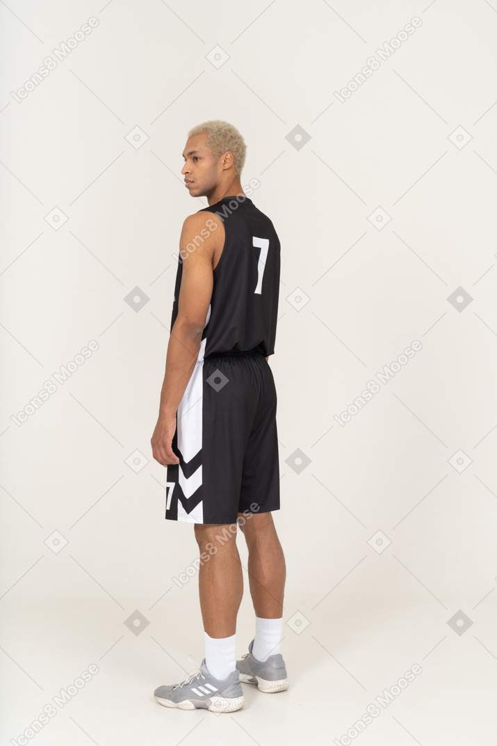 Vista posterior de tres cuartos de un joven jugador de baloncesto de pie y mirando a un lado