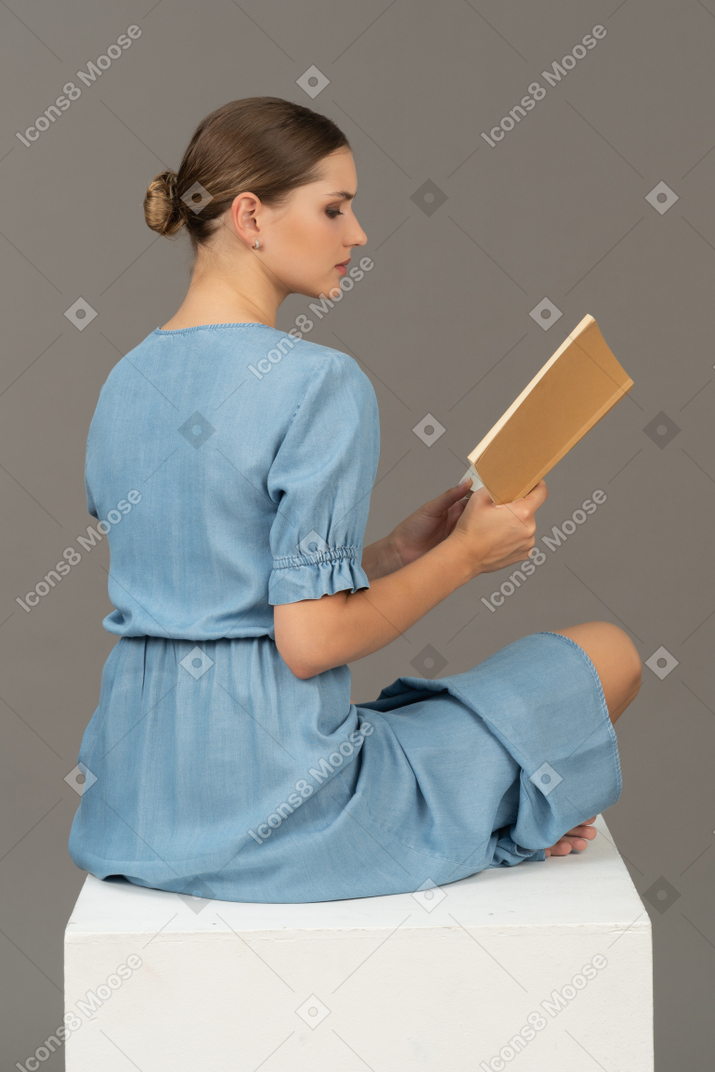 Side-back view of young woman sitting on a cube and reading book
