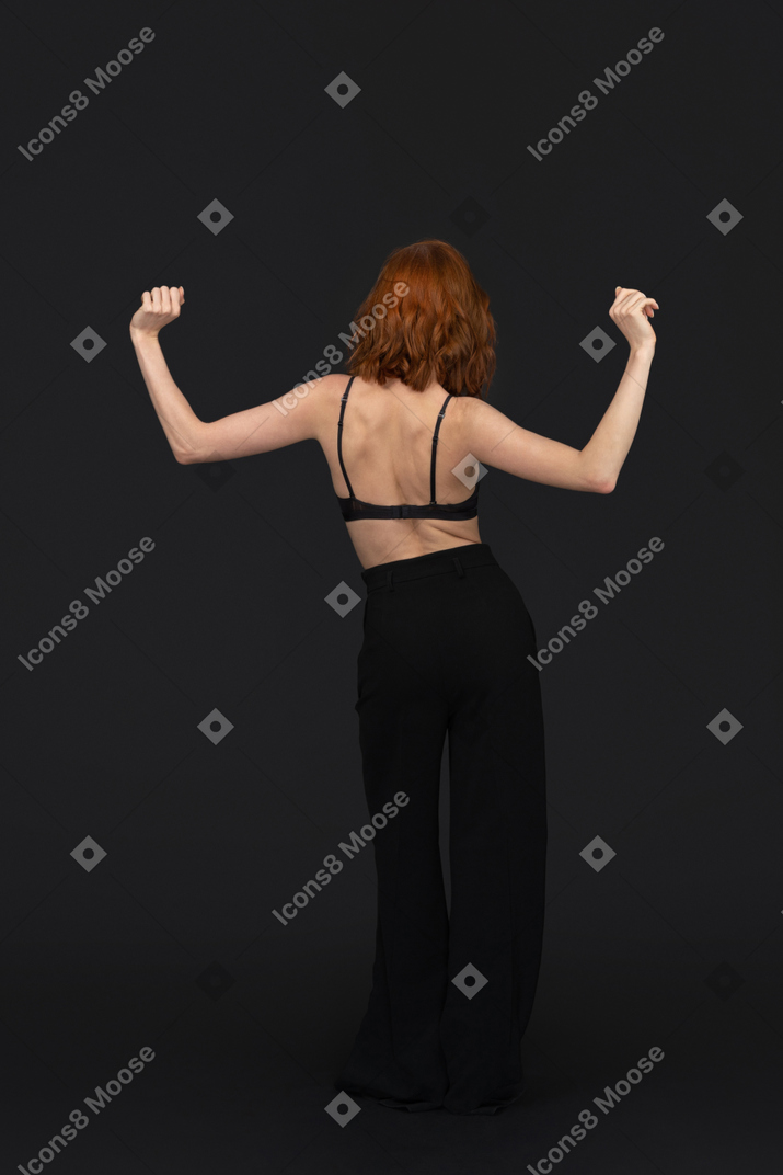 A back side view of the cute young woman dancing on the black background holding her fists up