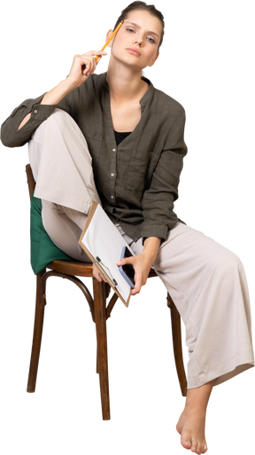 Front view of a thoughtful young woman wearing home clothes sitting on a chair and making notes
