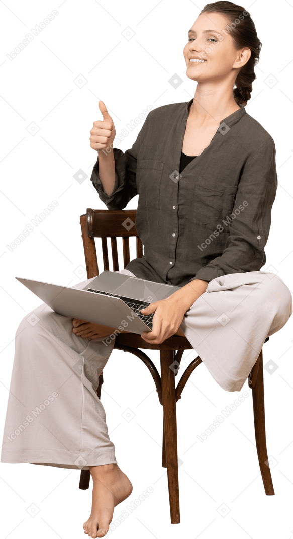 ノートパソコンで椅子に座って親指を上げて笑顔の若い女性の正面図