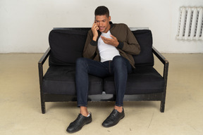 Front view of a perplexed young man sitting on a sofa and talking on his phone